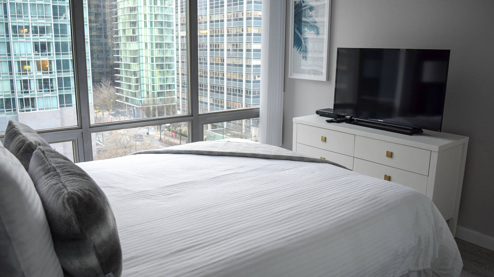 Fully furnished bedroom with white linens on a bed, white dresser and a flat screen TV and with a view at Vancouver Extended Stay