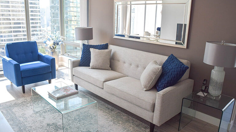 Living room with a light grey sofa, a blue chair and a glass coffee table in a move-in ready, fully furnished one bedroom and den apartment at Vancouver Extended Stay