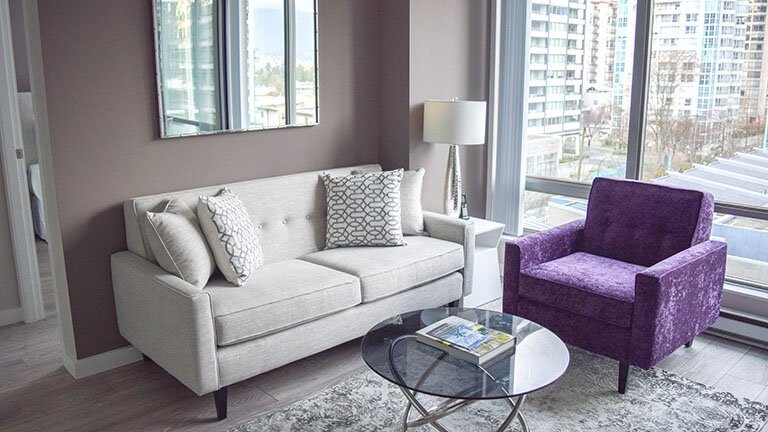 Living room with a light grey sofa, a purple chair and a round glass coffee table in a move-in ready, fully furnished one bedroom apartment at Vancouver Extended Stay